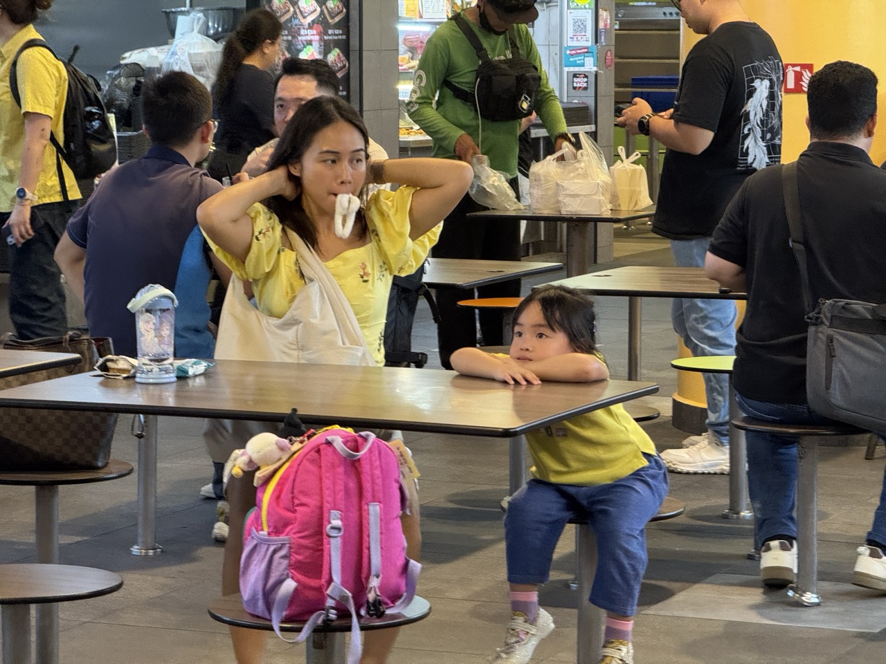 Mother and daughter seated at One Punggol Hawker Centre as customers and delivery riders collect food.